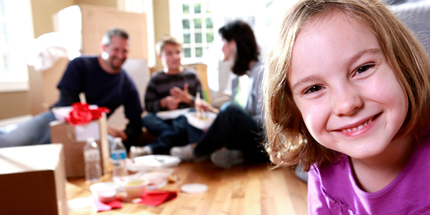 a young smiling girl close to the camera with adults sitting on a wooden floor and talking in the background