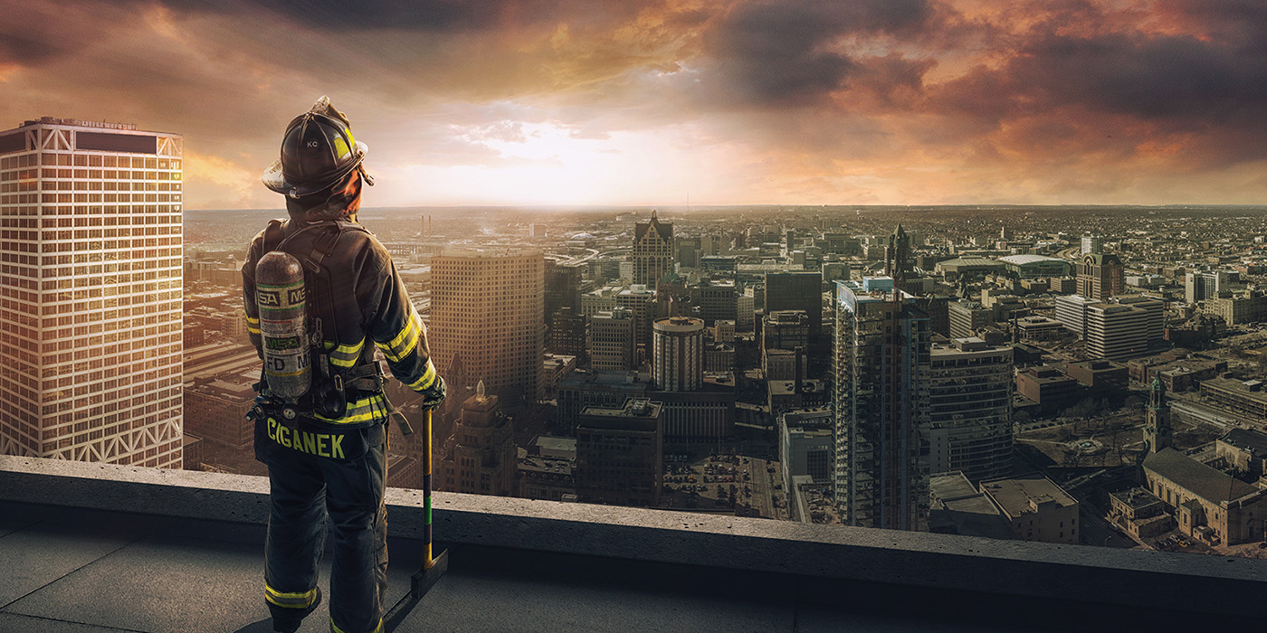 a firefighter in full turn-out gear stands watch from a rooftop looking out over a city with hundreds of buildings under a dramatic sky