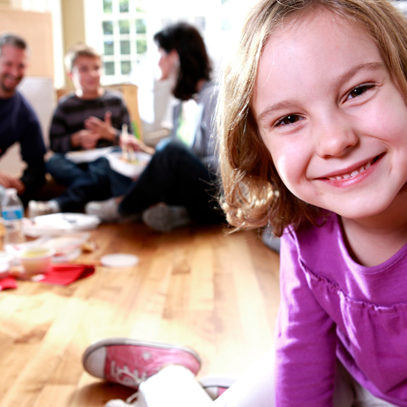 a young smiling girl close to the camera with adults sitting on a wooden floor and talking in the background